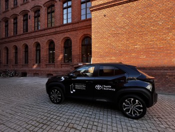 samochód Toyota Yaris Cross oklejony logotypem Toruńskiej Orkiestry Symfonicznej oraz Toyota Bednarscy na tle murowanego budynku